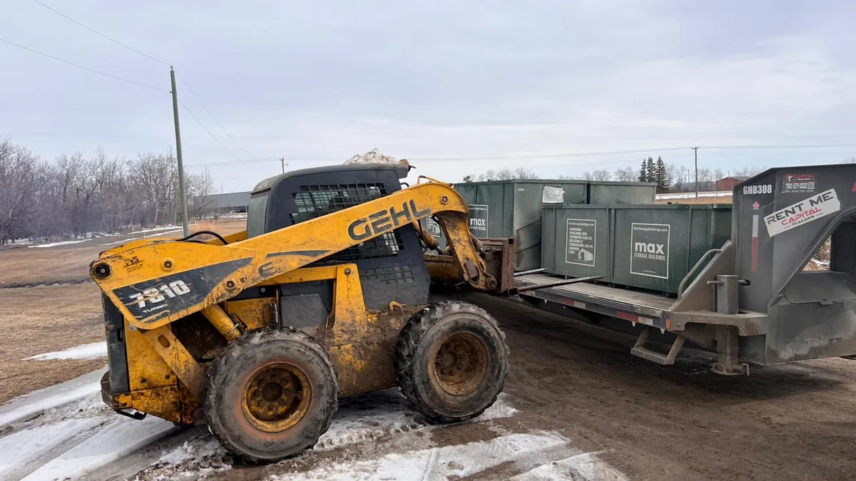 Skid steer beside crated building components on a flatbed trailer